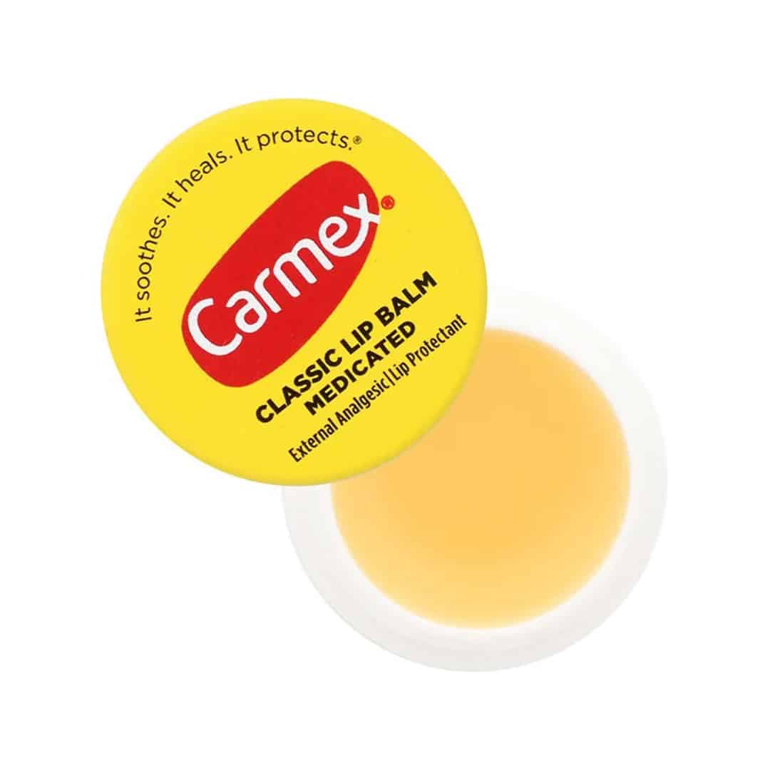 A yellow tin of Carmex Classic Medicated Lip Balm featuring the brand's red logo. Text on the lid reads: "It soothes. It heals. It protects.