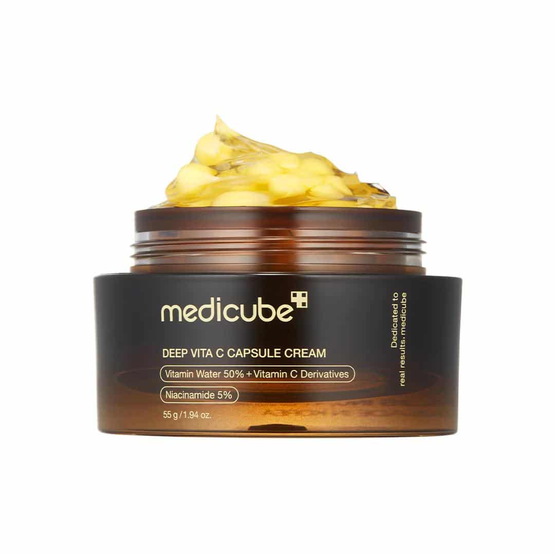 Open jar of Medicube Deep Vita C Capsule Cream 55 g on white background, showing translucent gel studded with golden vitamin C capsules alongside silver lid, illustrating dual-texture formula for brightening and hydration.